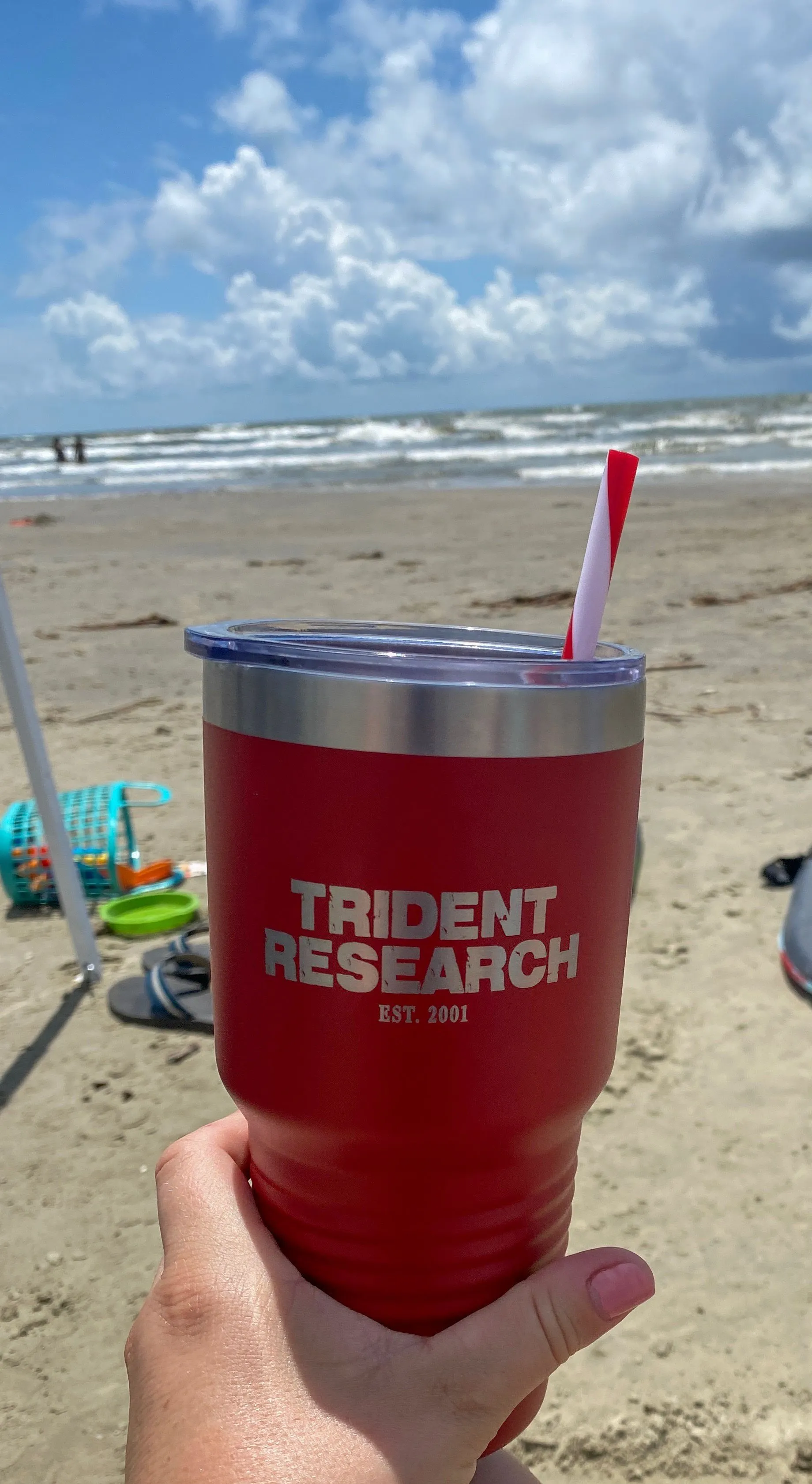 A hand holding up a red Trident Research tumbler on a sandy beach, waves and a blue sky behind it
