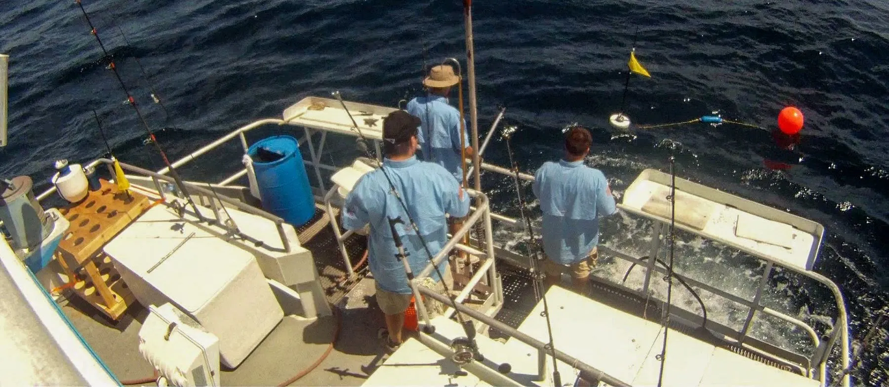 Seen from above, three crew in light blue shirts at the stern of a small boat watching a sensor float and an orange buoy on dark open water, a yellow marker flag beside them