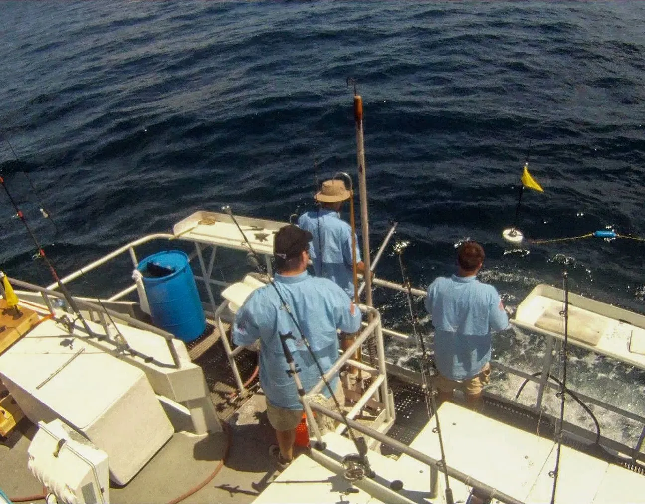 Seen from above, three crew in light blue shirts at the stern of a small boat handling a line that runs out to a yellow marker flag and floats on dark open water, fishing rods and a blue barrel beside them
