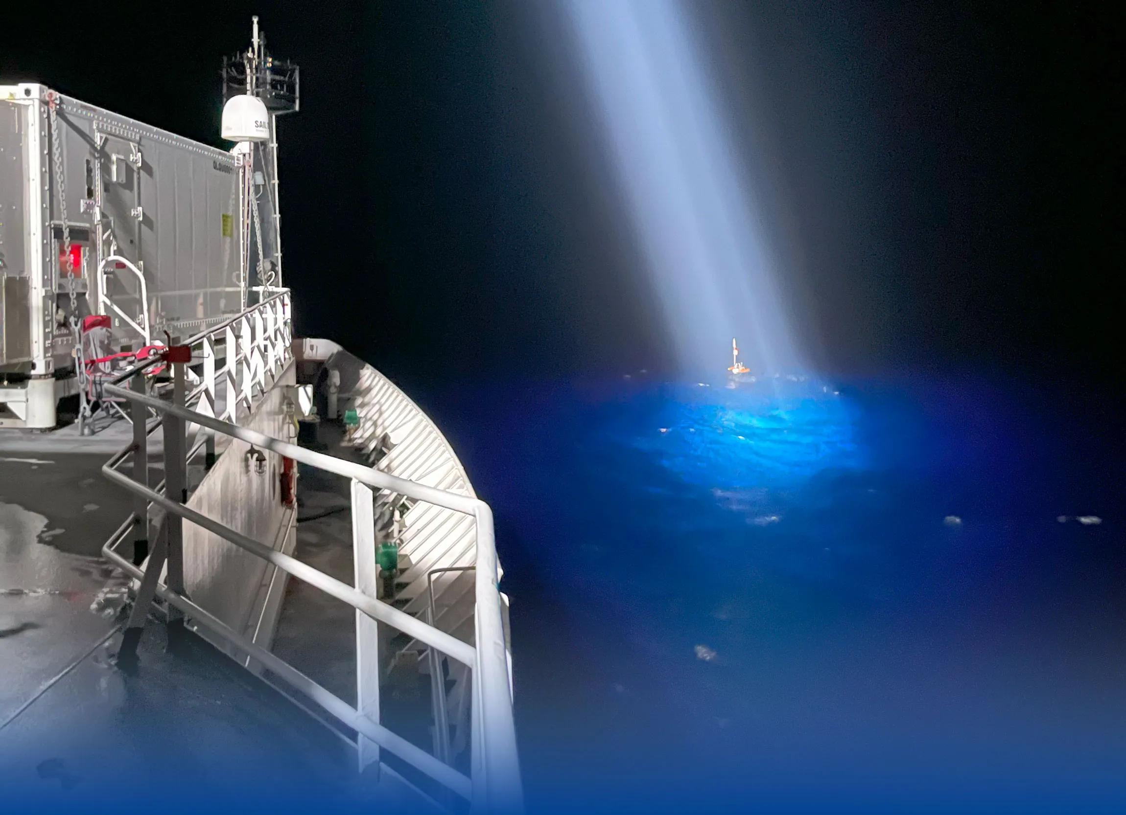 A Trident Research unmanned surface vehicle lit by a ship's searchlight during a night operation at sea