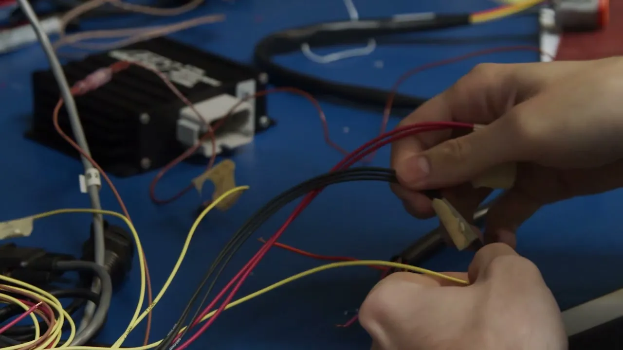 Two hands joining red and yellow wires to a connector on a blue workbench, beside a black electronics module with its harness plugged in