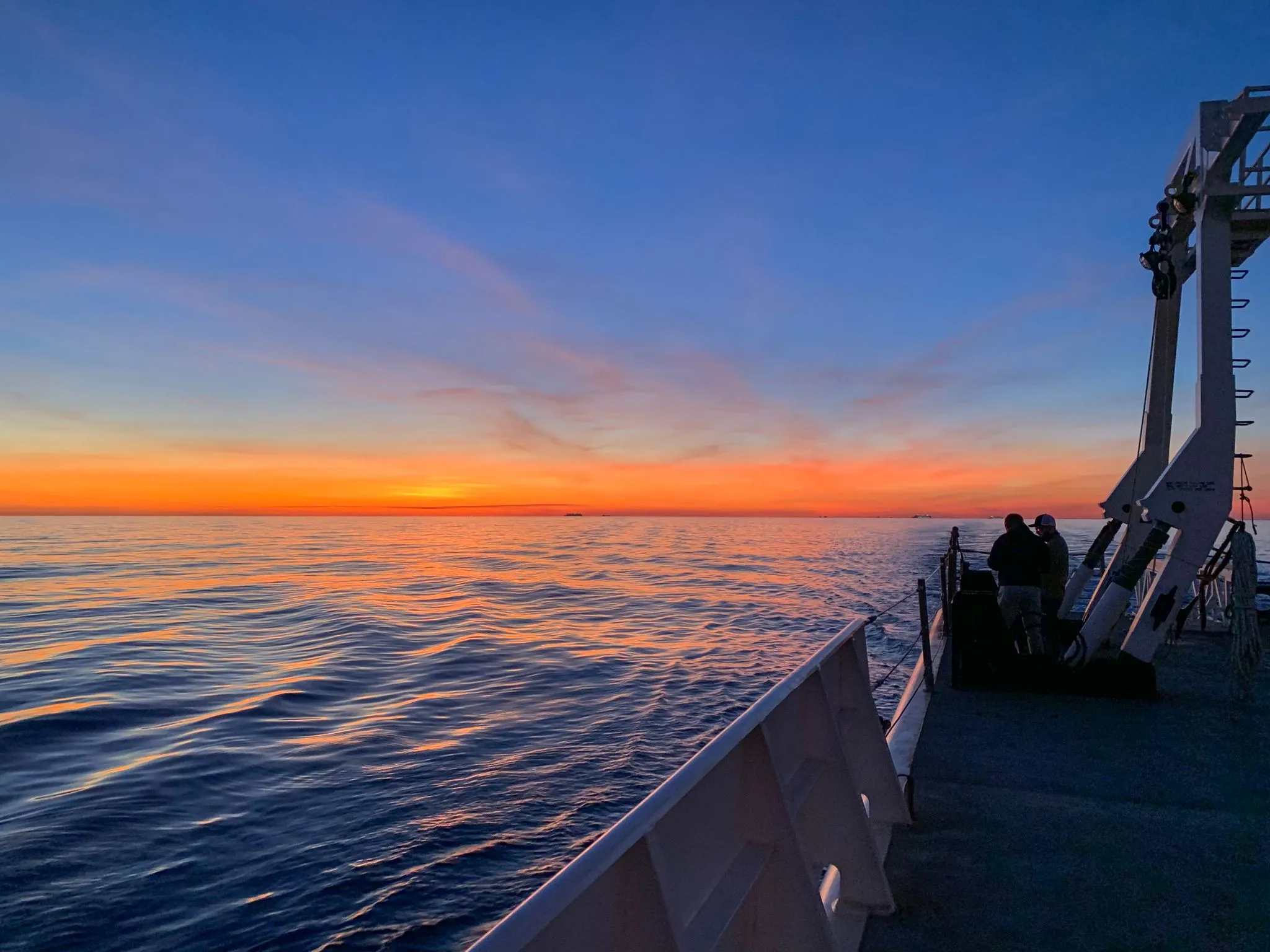 Two crew members silhouetted against an orange sunrise on the deck of a range support vessel, beside an A-frame crane