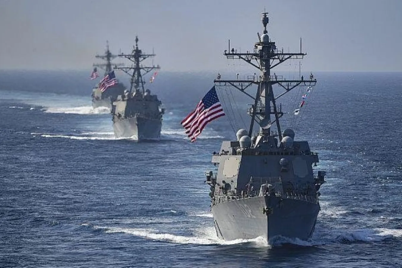 Three U.S. Navy destroyers sailing in column across open blue water, each flying a large American flag