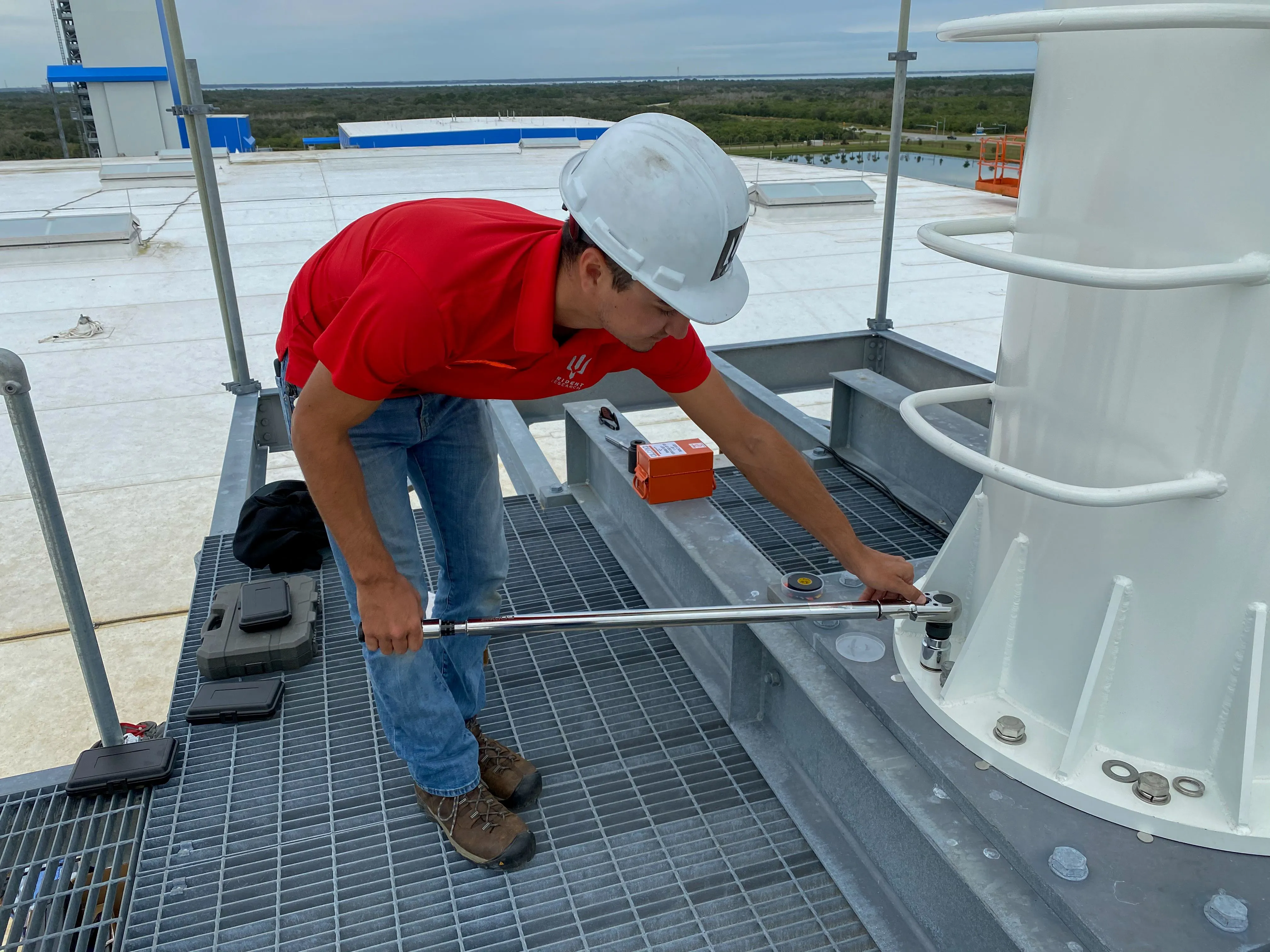 A Trident Research technician in a red company polo and white hard hat torquing hardware on a rooftop platform at a coastal launch complex