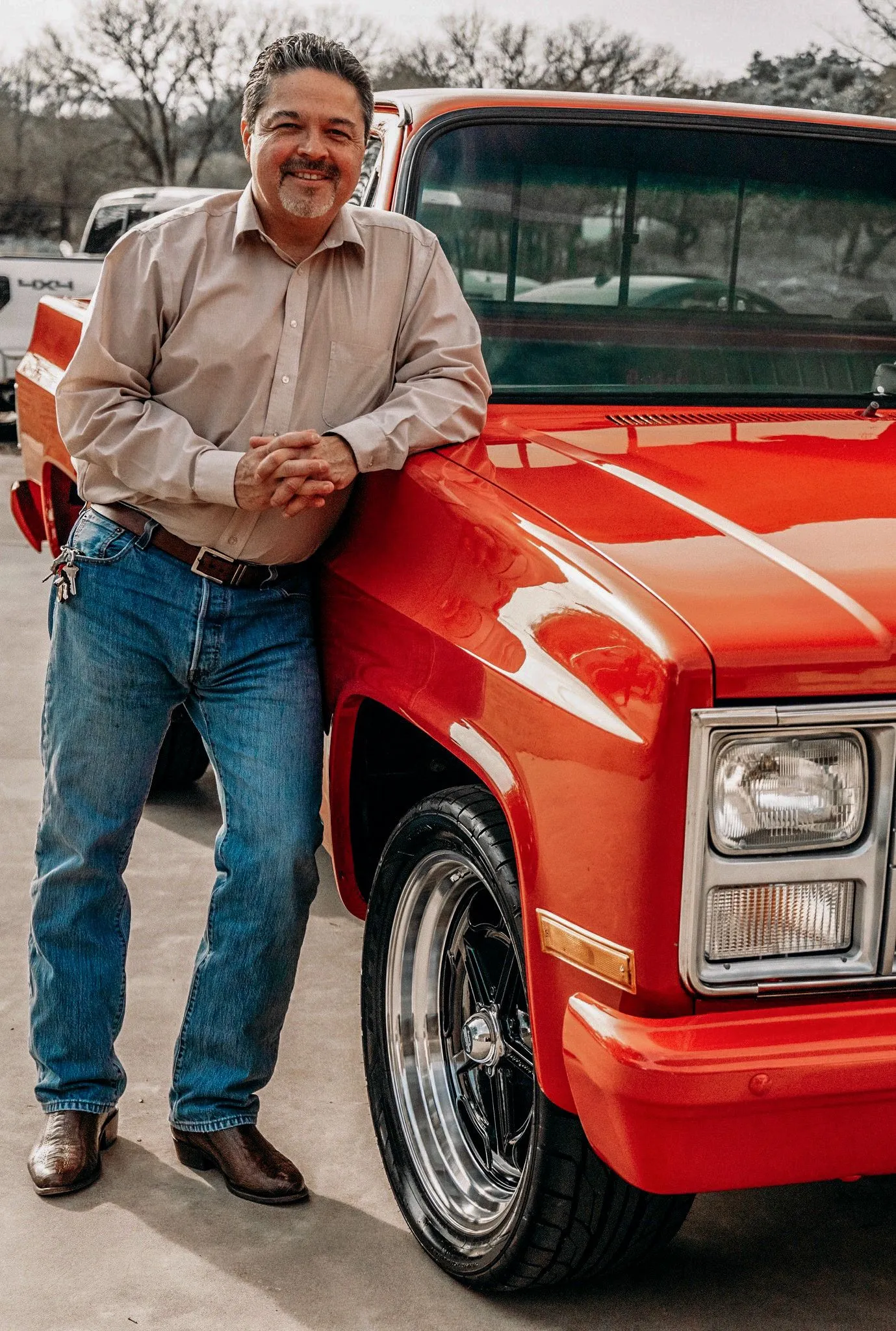 Trident Research's founder and president in a tan button-down shirt and jeans, leaning on the fender of a restored orange-red pickup truck, bare winter trees behind