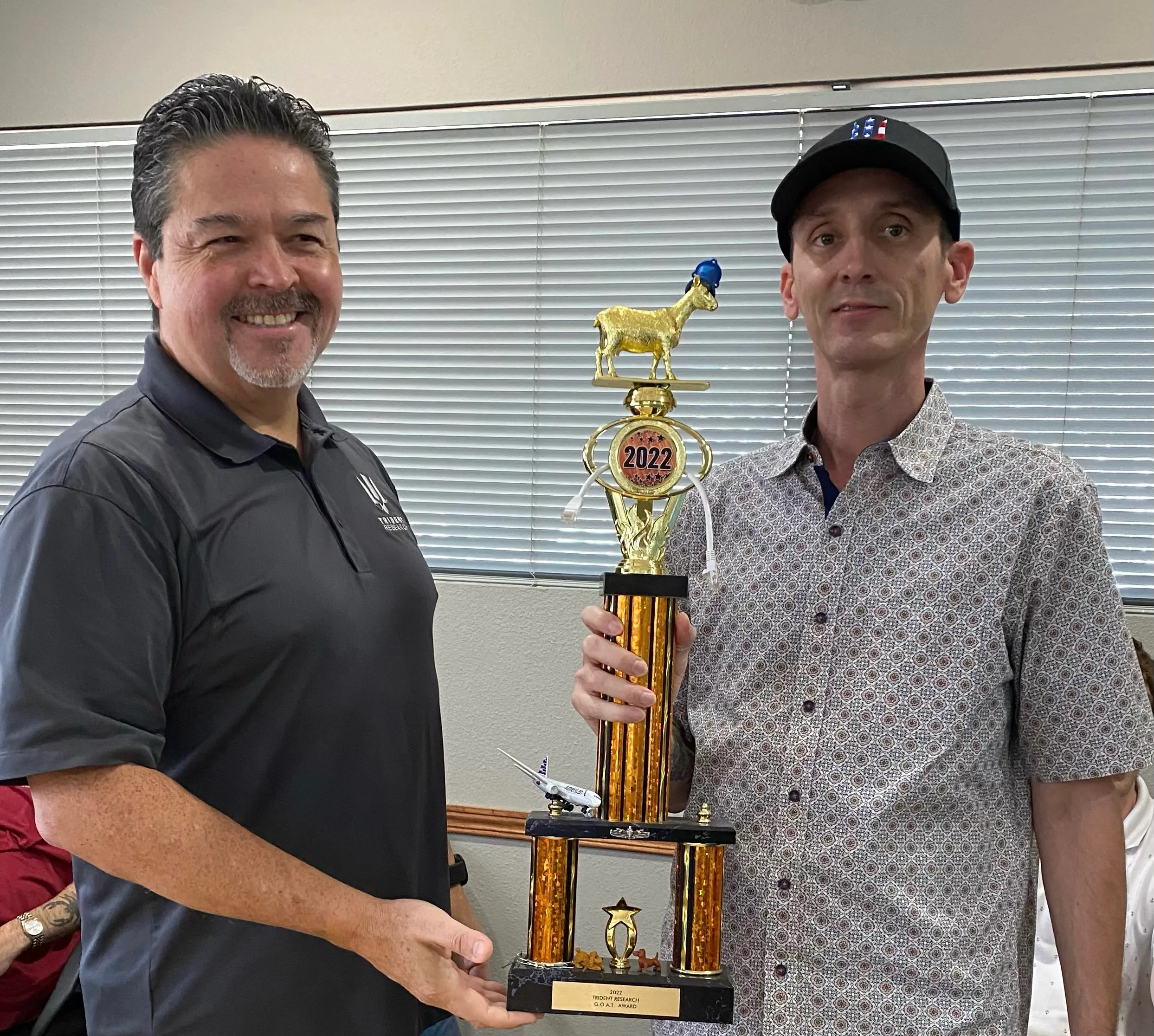 Two colleagues holding the 2022 Trident Research G.O.A.T. Award — a gold trophy topped with a goat figurine wearing a small blue hard hat
