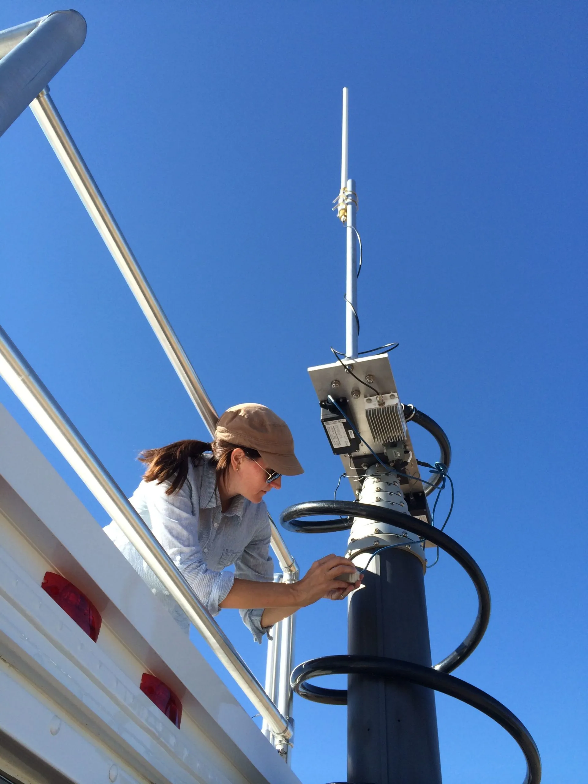 A woman in a tan cap and sunglasses leaning over the rail of a white truck bed to adjust a sensor unit mounted partway up a tall black mast with a whip antenna, against a clear blue sky