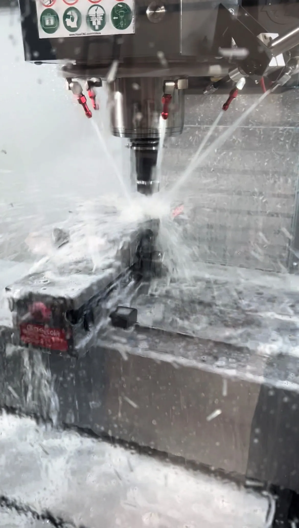Coolant spraying from the spindle of a Haas vertical mill onto a workpiece clamped in a vise, the splash frozen mid-cut