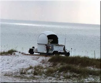 A white domed optical tracking unit on a trailer parked on a sandy rise beside the sea, with a person at its side