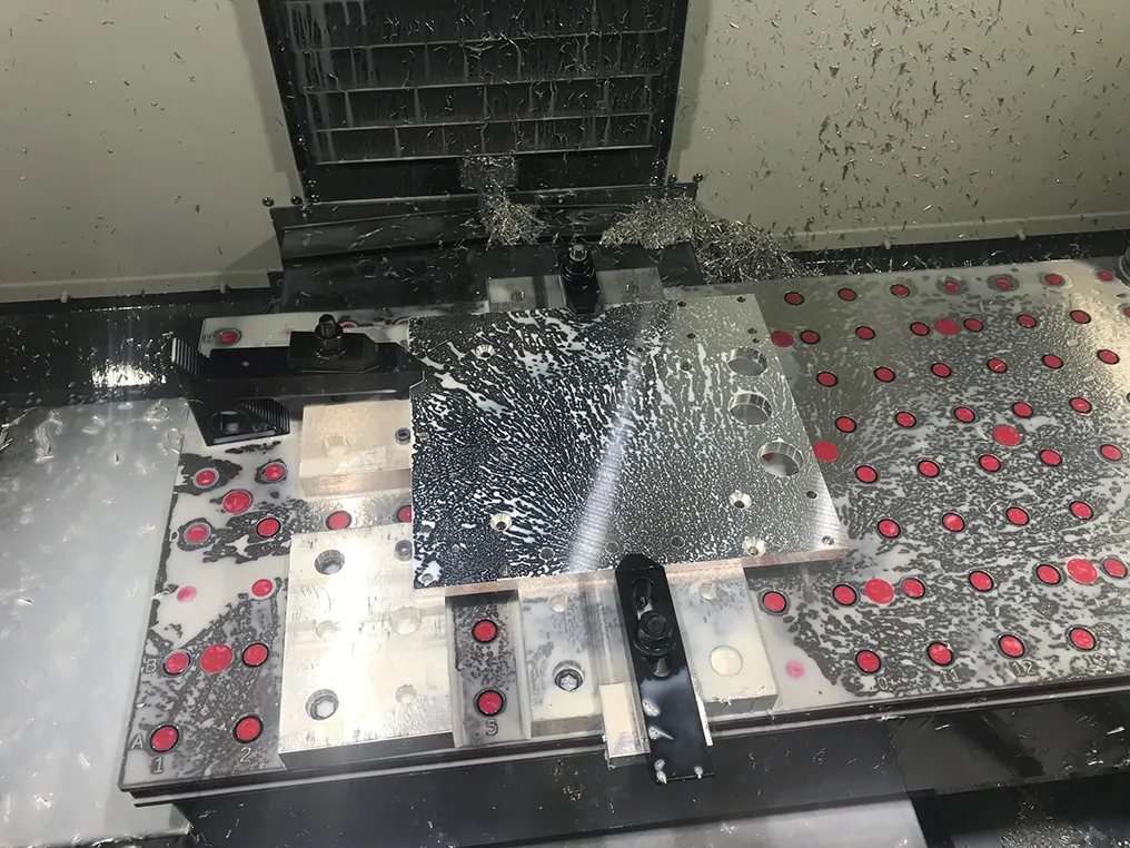 An aluminum plate clamped to a red-dotted fixture plate inside a mill, chips and coolant spread across it mid-job