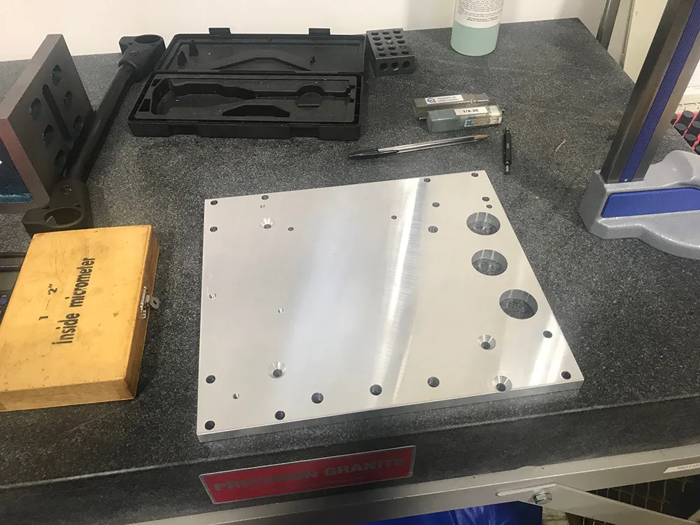The finished aluminum plate, its machined holes clean, resting on a granite surface plate beside a dial indicator case and an inside micrometer box
