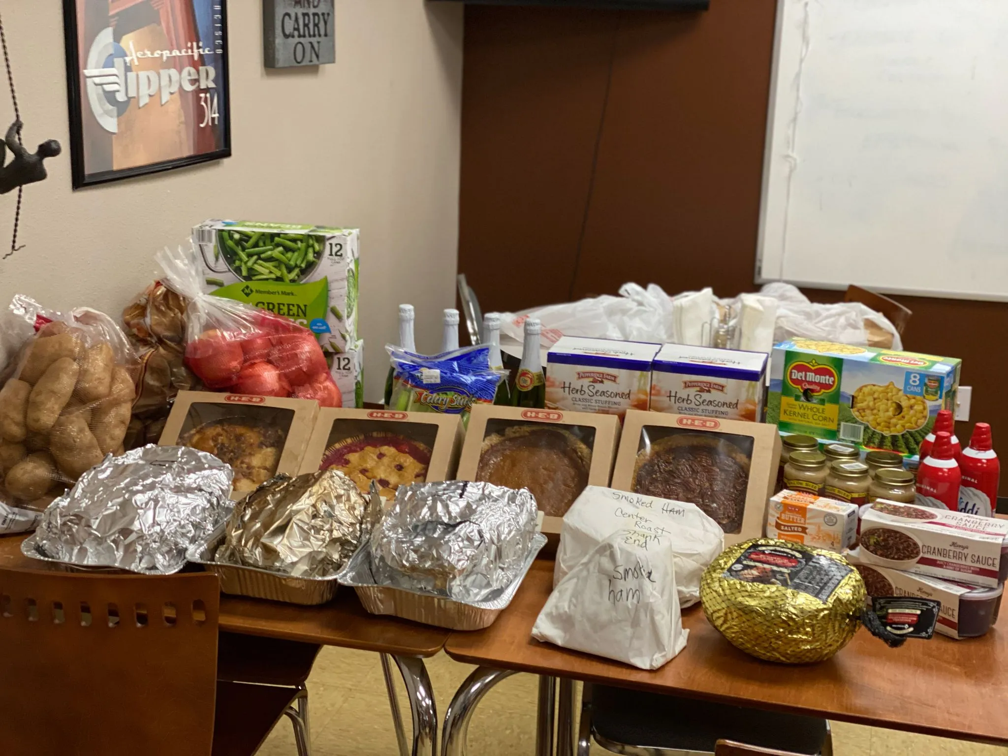 An office table loaded for a potluck: foil-covered trays, pies, potatoes and groceries under a framed print on the wall