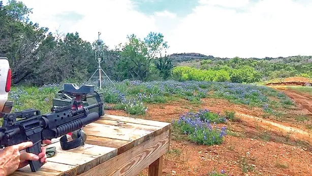 A rifle fitted with a grenade launcher rests on a wooden shooting bench among bluebonnet flowers, with a Trident sensor on a tripod standing in the scrubland and trees in the distance