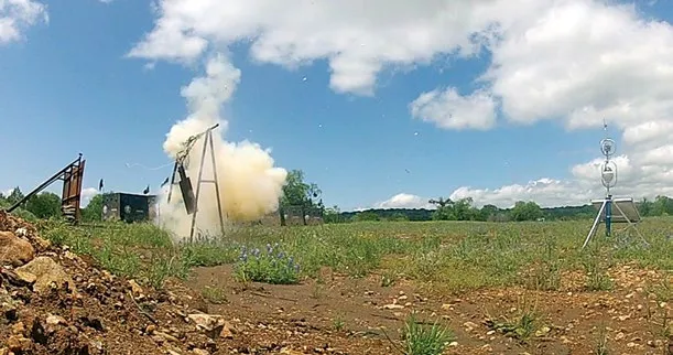 A white cloud of smoke and dust billows around a wooden target frame on a grassy range under a blue sky, with a Trident sensor on a tripod standing at the right