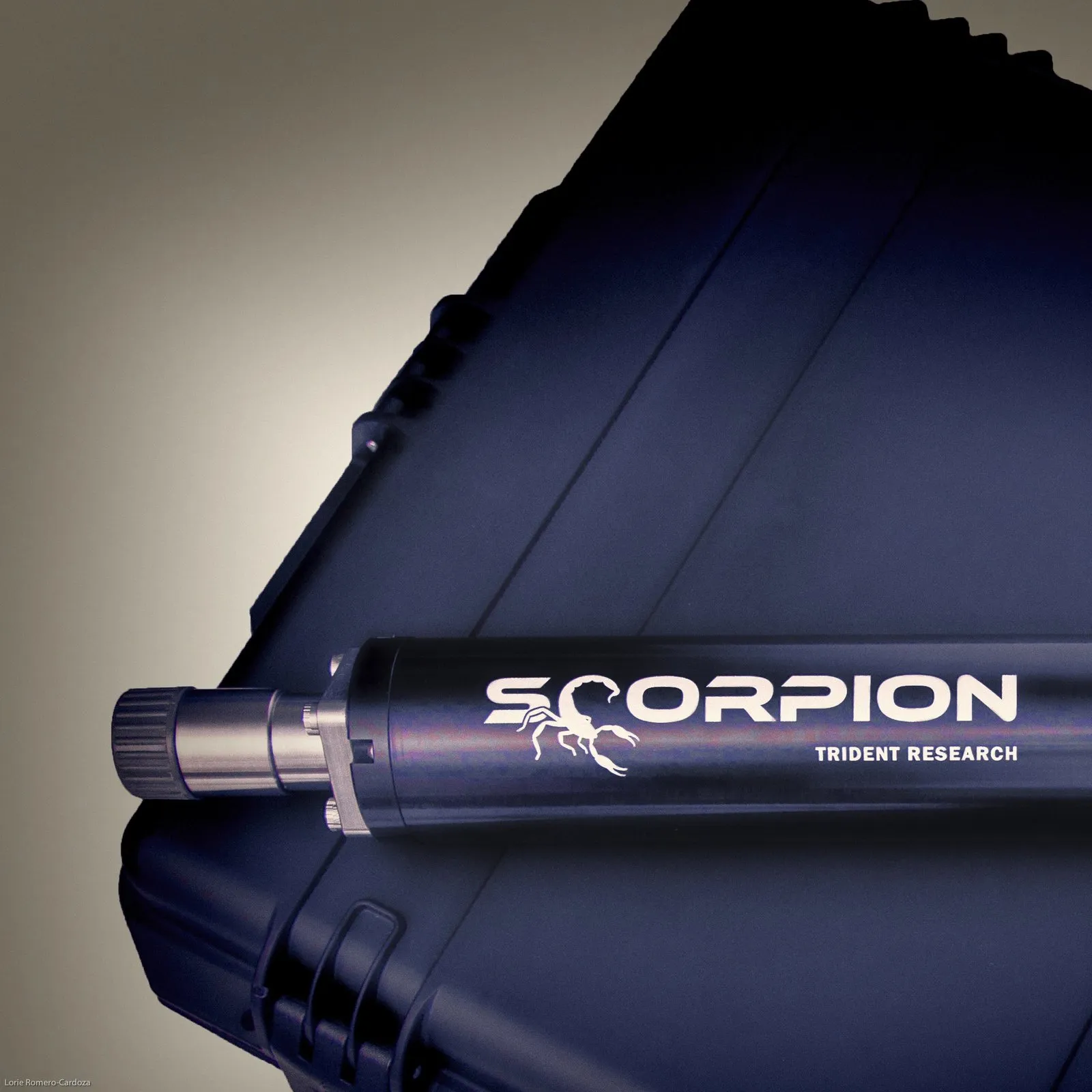 A close view of the black Scorpion sparker, its steel end fitting and knurled cap pointing left, with white SCORPION lettering, a scorpion emblem and TRIDENT RESEARCH along the tube, lying on a navy hard case against a grey-brown backdrop