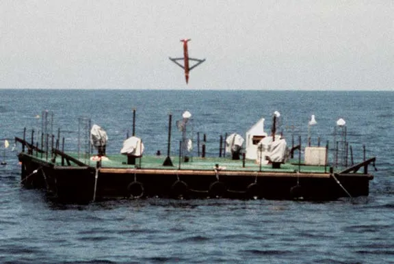 A Small Diameter Bomb, a small winged munition, falls nose-down above a flat floating target platform with equipment and instrument housings on its deck, on calm blue-grey water.