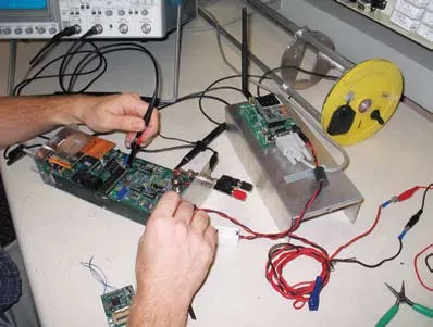 Two hands hold test probes over a circuit board on a laboratory bench, with a second board, cables and an oscilloscope behind it.