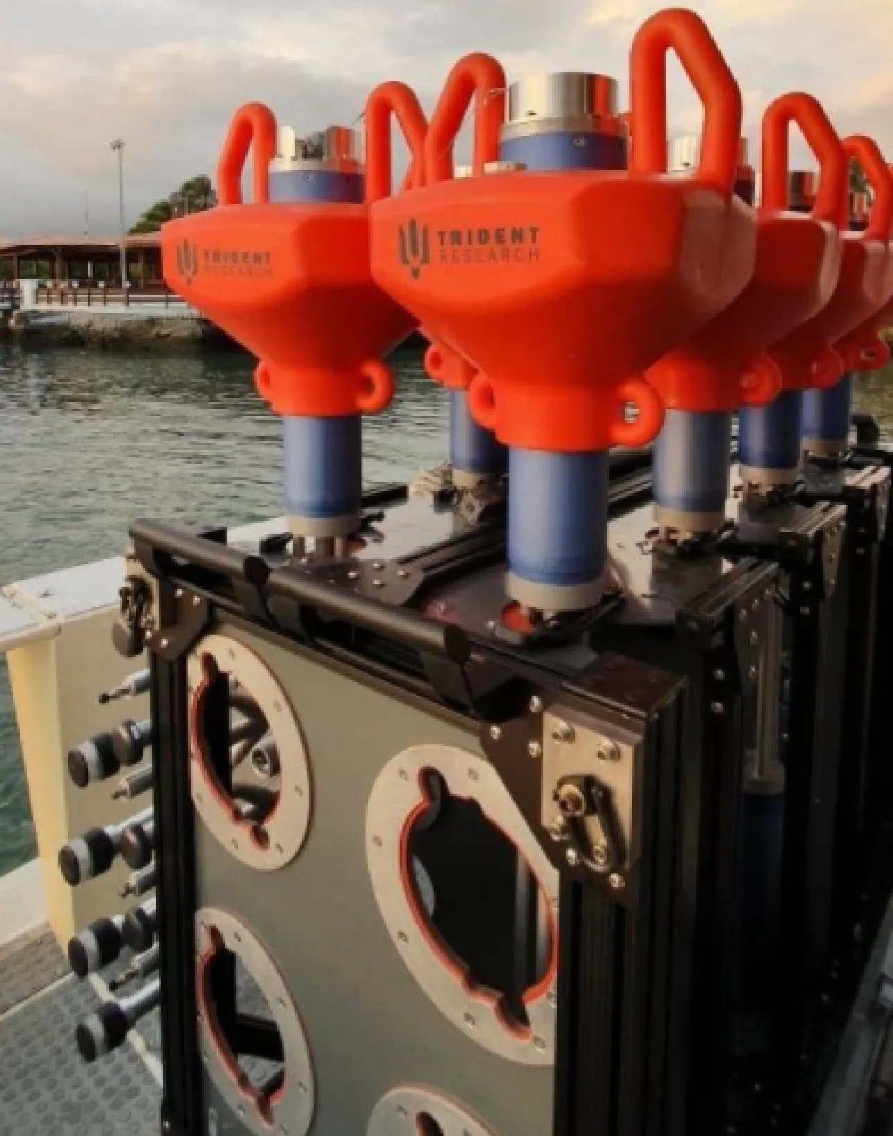A row of orange Trident Research sensor pods with blue cylindrical bodies mounted on a black aluminum-framed enclosure on a boat deck, with calm water and a pier behind