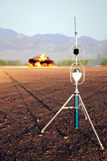 A Trident acoustic sensor on a white tripod, topped with a ring antenna and a mast, standing on dry red-brown ground in the foreground while a fireball and smoke from a detonation burst on the flat range behind it, with hills under a blue sky beyond