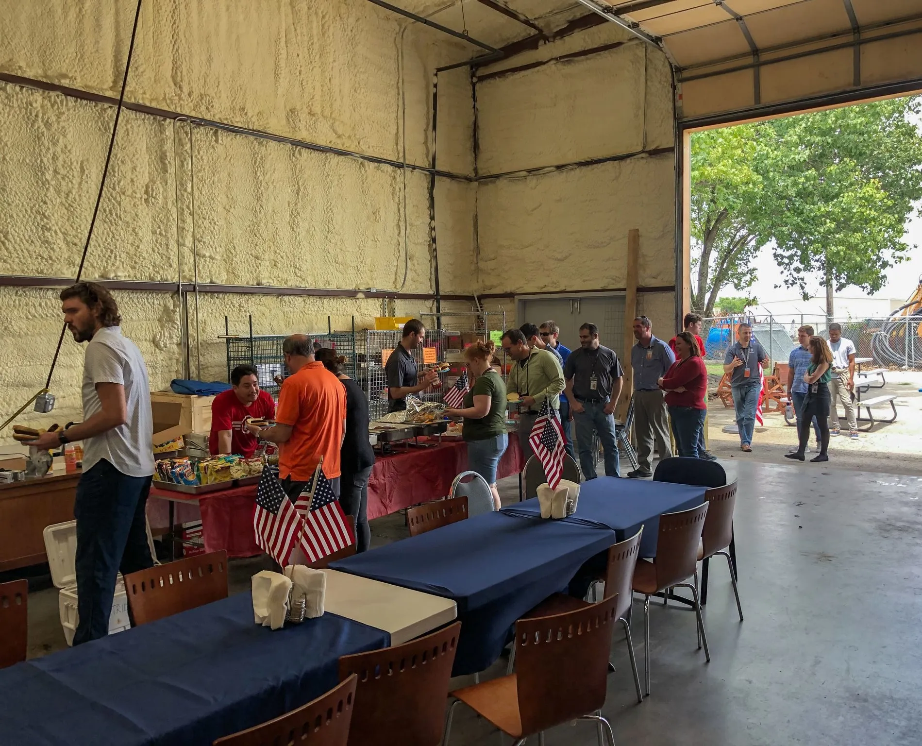 Colleagues lining up for a cookout inside the Trident shop bay, small US flags on red and navy tables and the roll-up door open to the trees outside