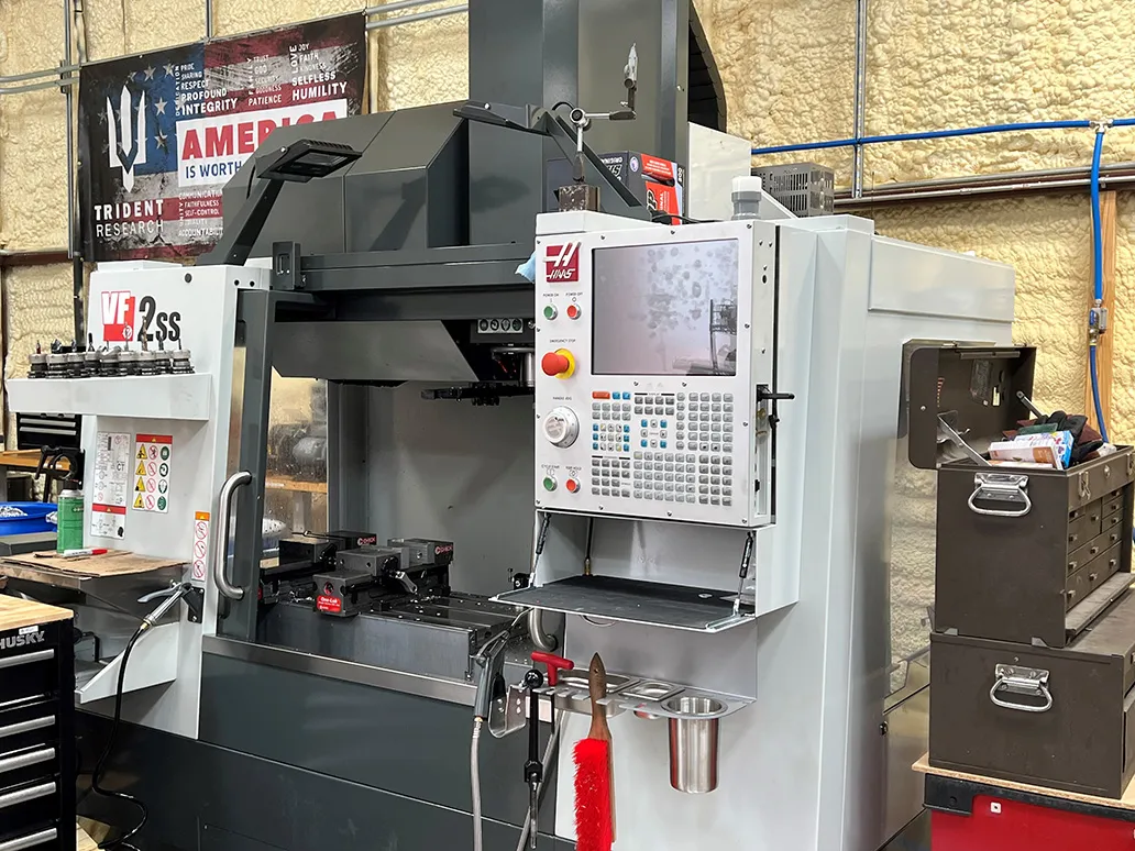 A Haas VF-2SS vertical mill in the Trident machine shop, a Trident Research "America is worth it" banner on the insulated wall behind it