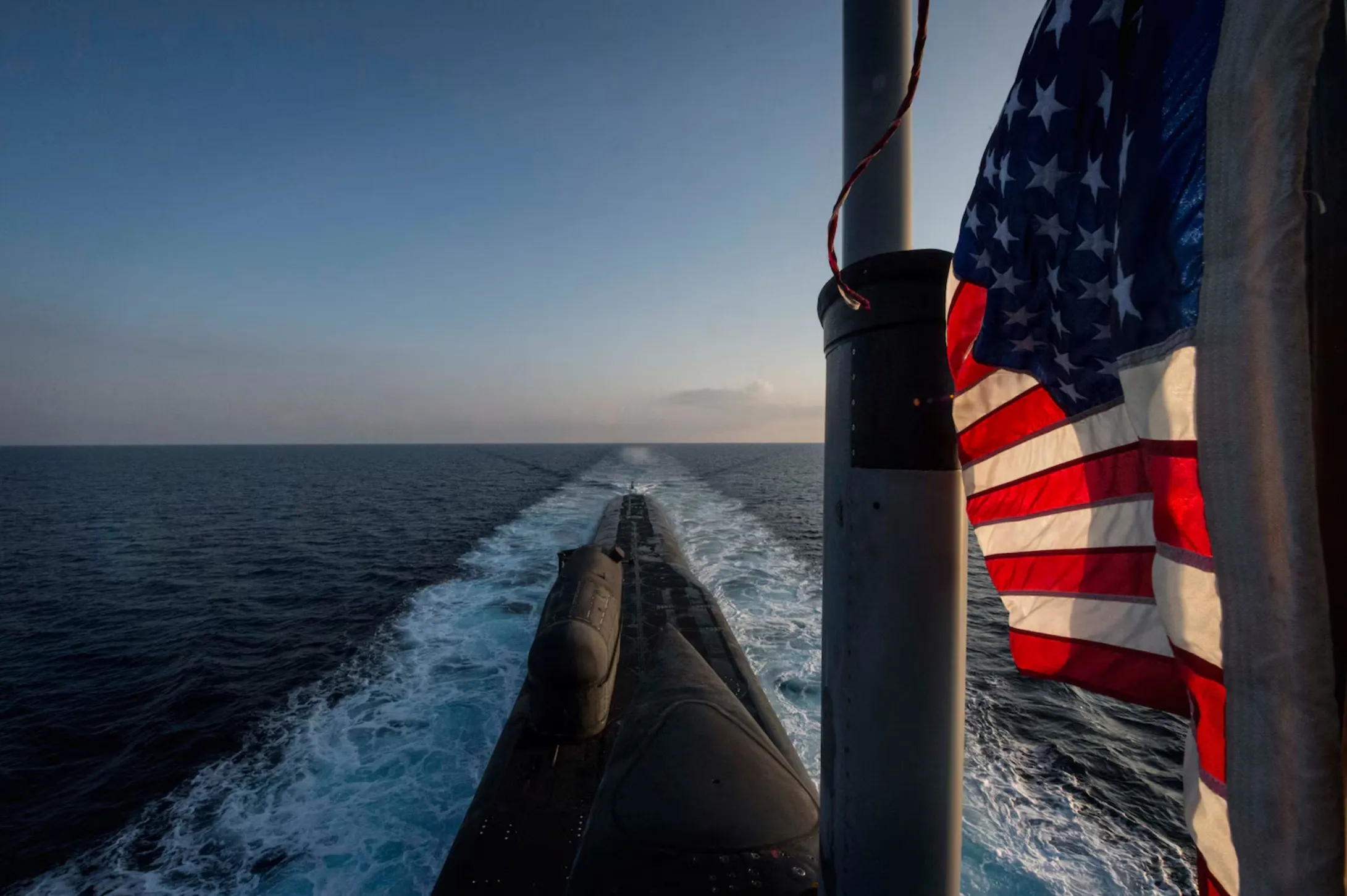 Looking aft along a surfaced U.S. Navy submarine at dusk, its wake spreading behind it and an American flag flying from a mast in the foreground