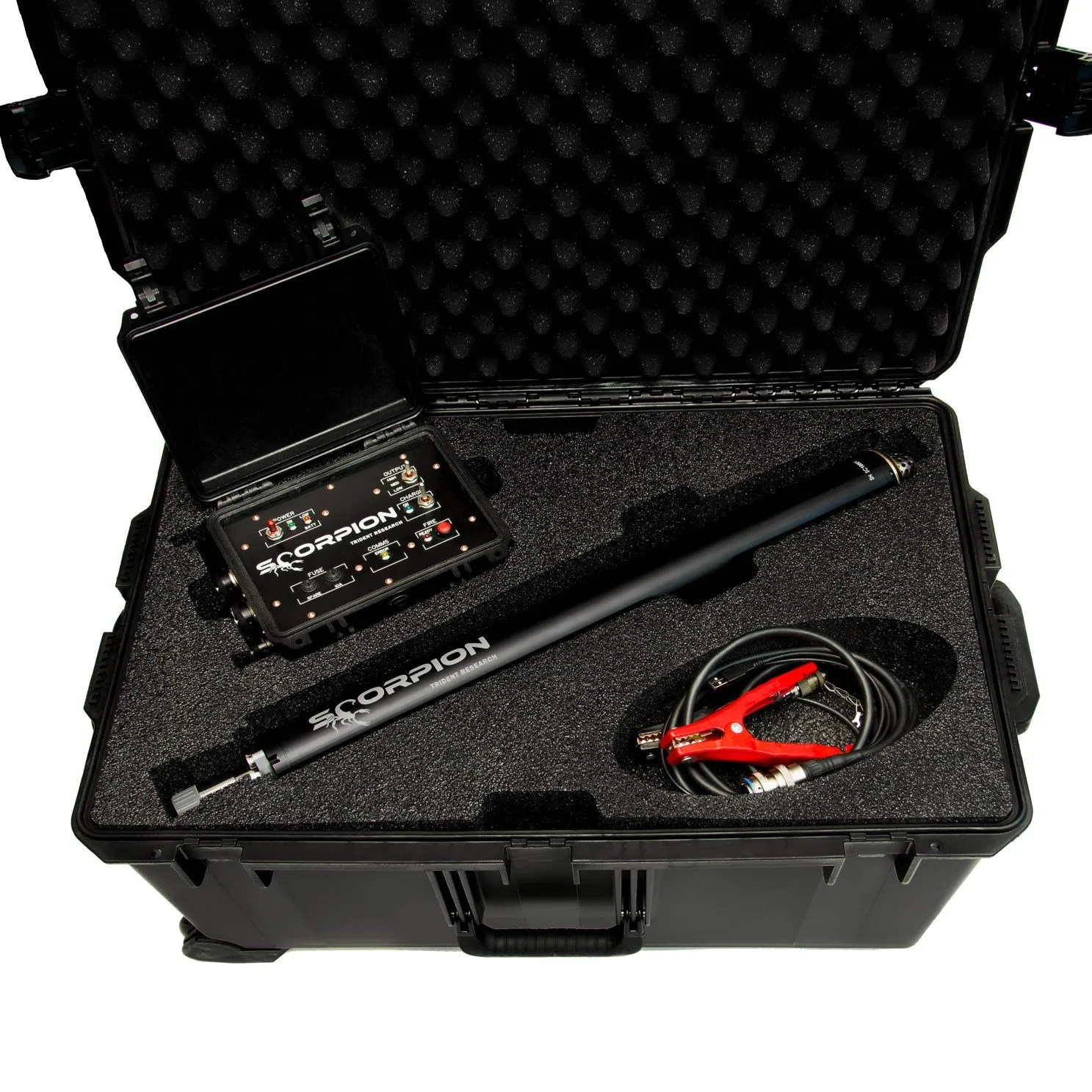 A black wheeled hard case opened from above: in grey foam, a control unit with its own lid raised at upper left, the black Scorpion sparker tube lying diagonally across the middle, and a coil of lead with red battery clamps at lower right