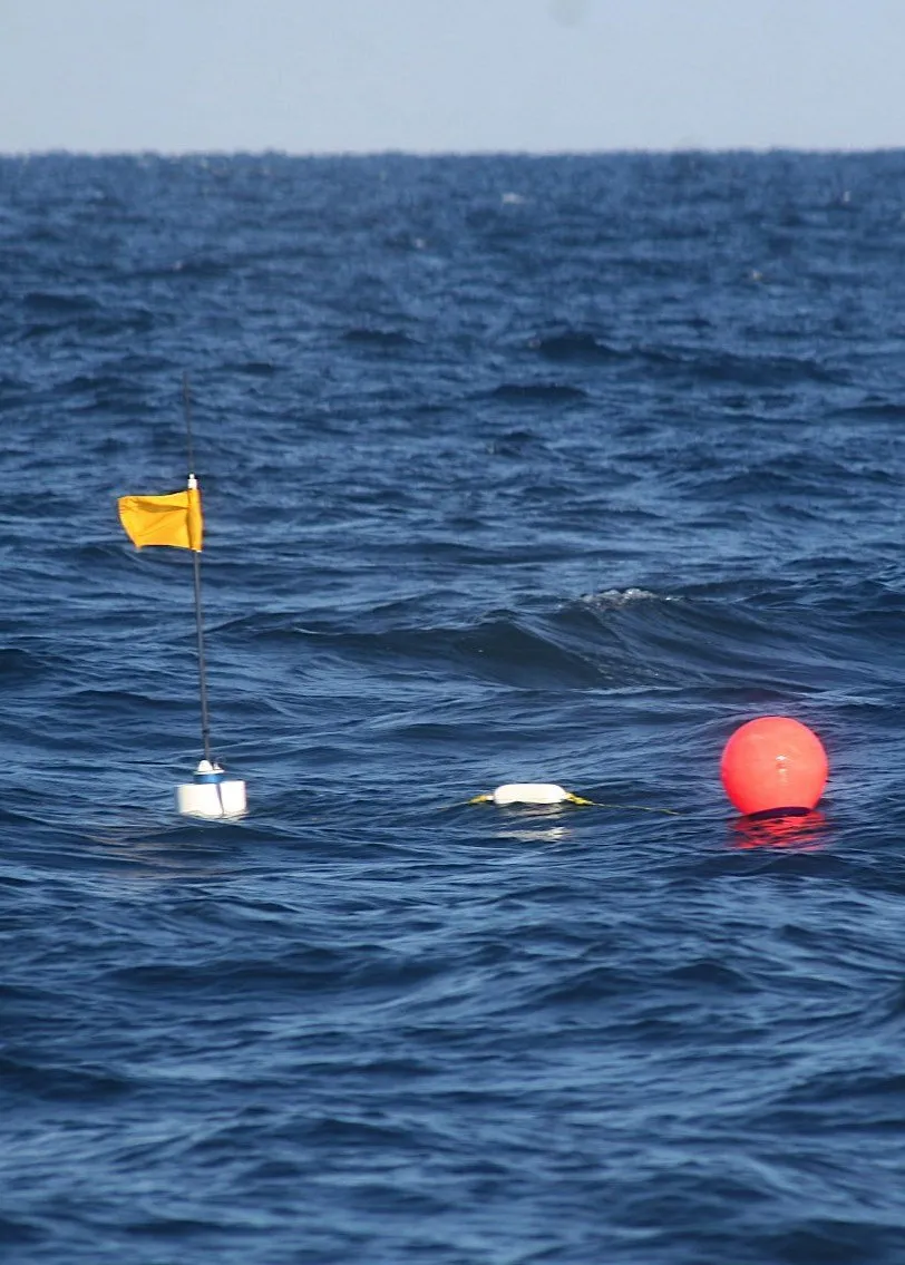 Three sensor floats on dark blue open water, one with a yellow flag on a mast, one a white float and one a red-orange buoy