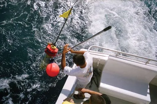 Seen from above, a crew member on the deck of a small boat lowers a TARGT sensor with a long pole into churning water; a red buoy floats beside it.