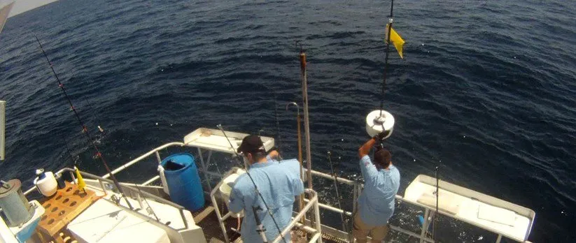 Two men in light blue shirts on the deck of a boat, one lowering a white sensor float on a line over the rail, with dark open water below and a yellow flag on a pole