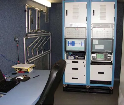 The inside of the instrumentation shelter: two blue equipment racks with monitors, a desk with a laptop and an office chair, and sensor mounting brackets on the wall