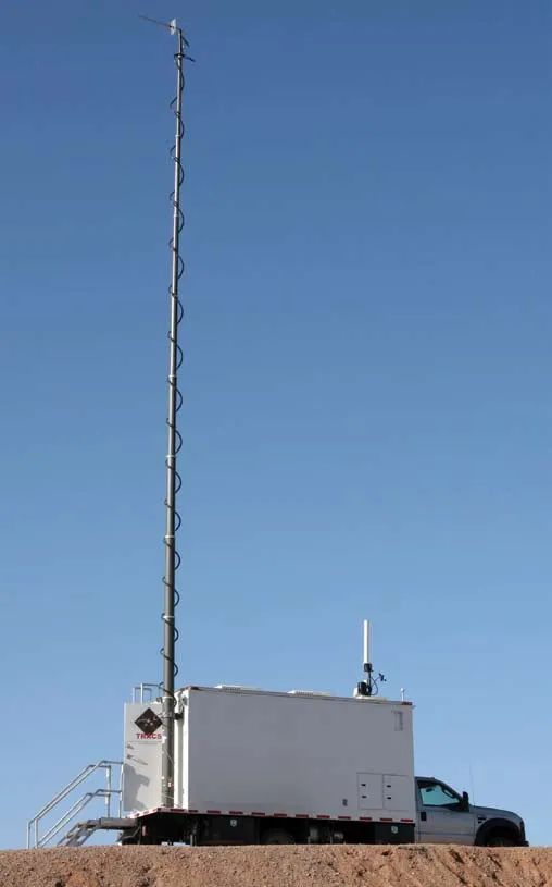 A white truck-mounted instrumentation shelter marked TRACS with a tall extended mast, standing on a gravel rise under a clear blue sky