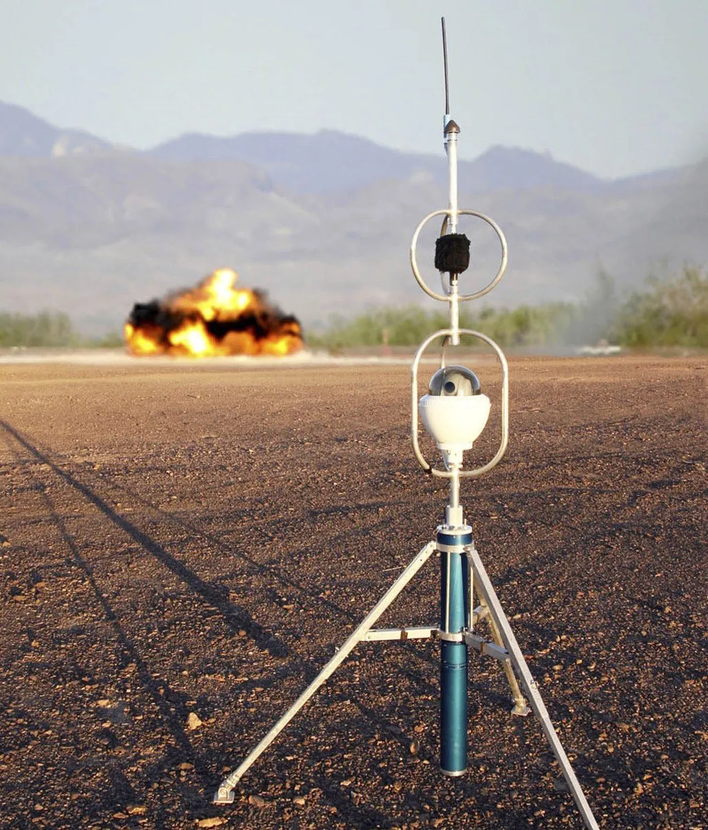 A sensor on a tall tripod with two rings and a whip antenna standing on a gravel desert flat, with a fireball from a detonation on the ground behind it and mountains in the distance