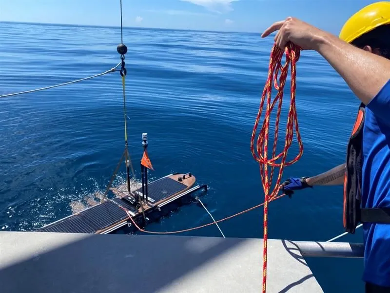 Seen from a ship's rail, a crew member in a yellow hard hat and orange life vest holds a coiled red-and-yellow line while a small unmanned surface vehicle with an orange flag hangs from a crane strap at the surface of calm blue water