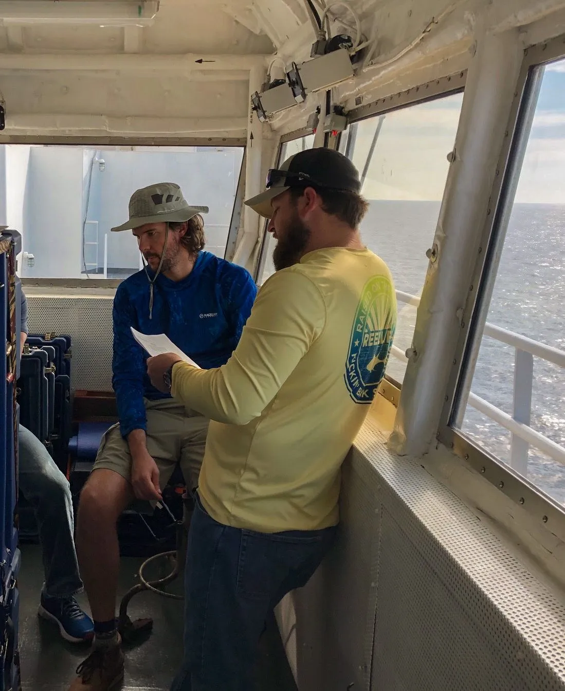 Two crew in a ship's wheelhouse reading over a sheet of paper together, open sea through the windows